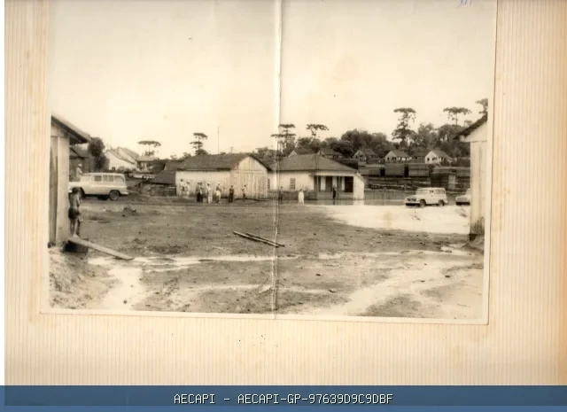 Área urbana antiga não pavimentada, com construções, veículos, pessoas e araucárias ao fundo.