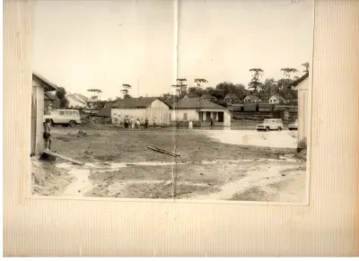 Área urbana antiga não pavimentada, com construções, veículos, pessoas e araucárias ao fundo.