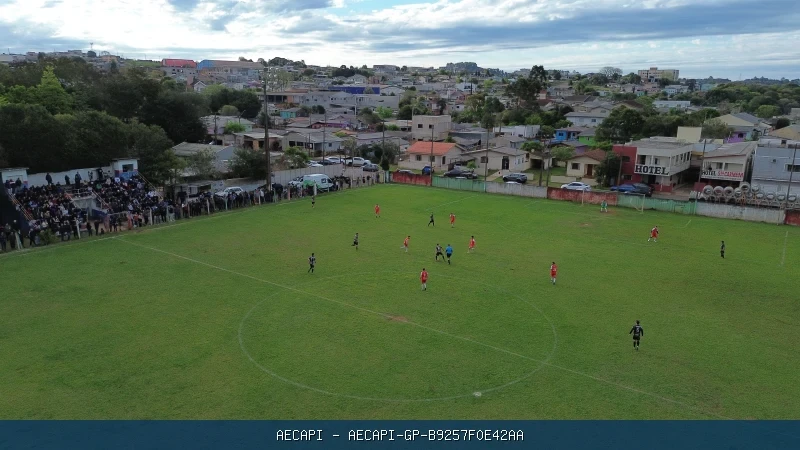 Vista aérea de partida de futebol em campo gramado Sertanejo com público ao redor e área urbana ao fundo.
