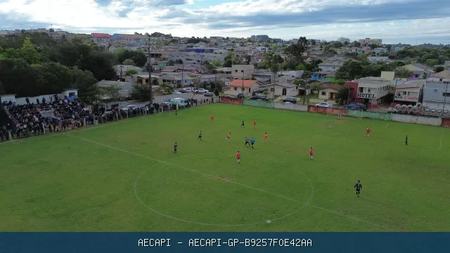 Vista aérea de partida de futebol em campo gramado Sertanejo com público ao redor e área urbana ao fundo.