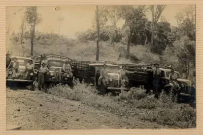 Fileira de caminhões antigos com pessoas ao lado, em uma área rural não pavimentada.