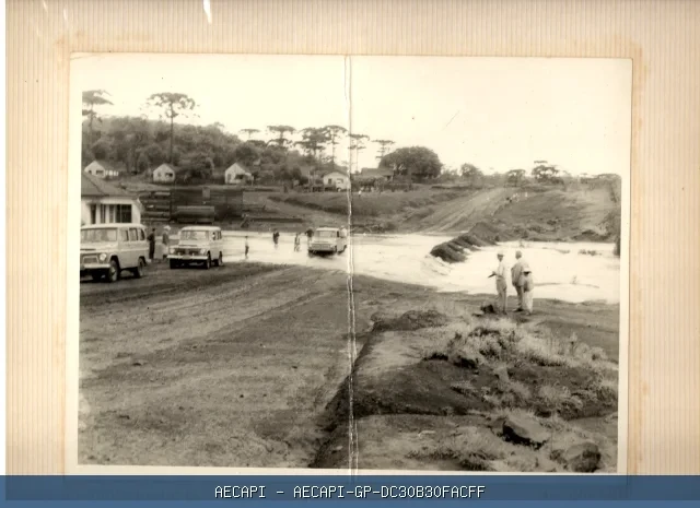Via antiga não pavimentada com veículos, pedestres, casas e araucárias ao fundo.