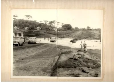 Via antiga não pavimentada com veículos, pedestres, casas e araucárias ao fundo.